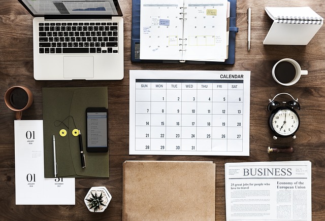 Top view of a desk highlighting a desktop calendar and agenda, as well as a clock and a laptop. Other standard items for a desk are neatly set nearby.